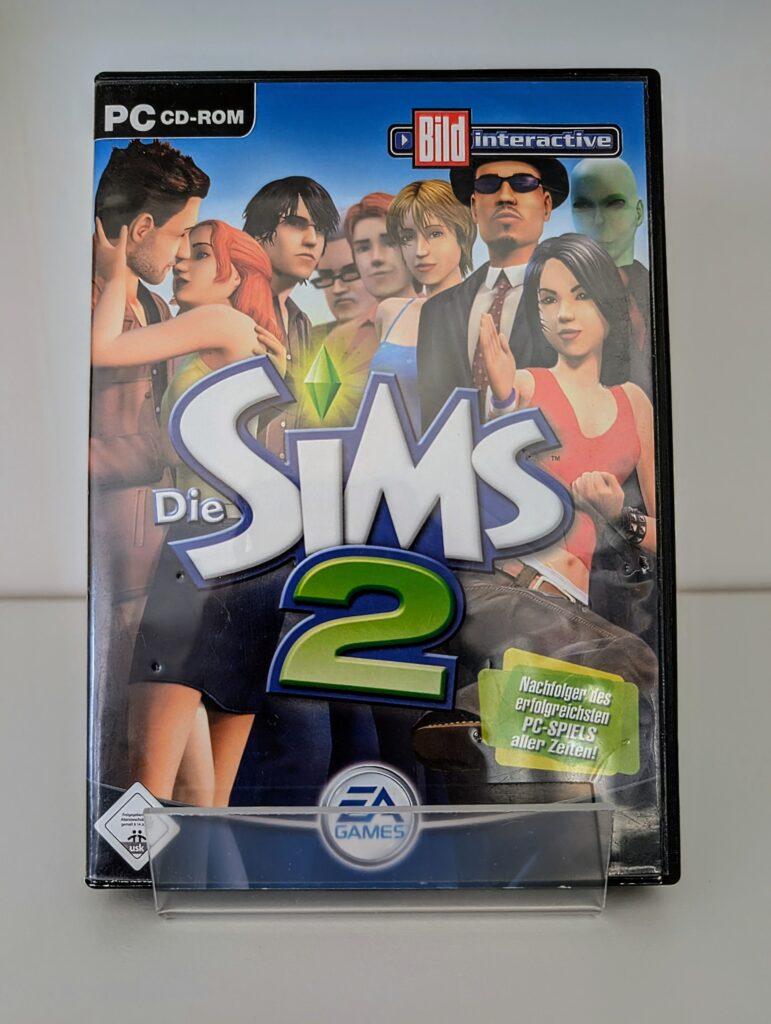Das Foto zeigt die Verpackung der CD-ROM des Basisspiels Die Sims 2. Darauf sind verschiedene Spielfiguren zu sehen.